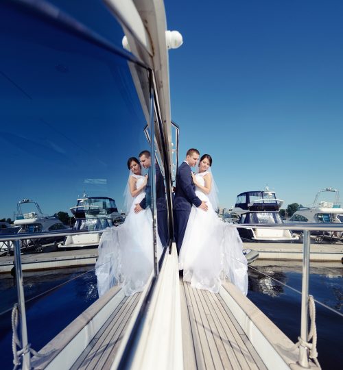 Wedding couple is hugging on a yacht. Beauty bride with groom. Beautiful model girl in white dress. Man in suit. Female and male portrait. Woman with lace veil. Cute lady and guy outdoors